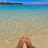 Feet resting in shallow clear water on a tropical beach with palm trees and boats in the distance.