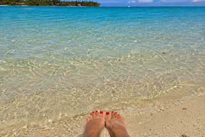 Feet resting in shallow clear water on a tropical beach with palm trees and boats in the distance.