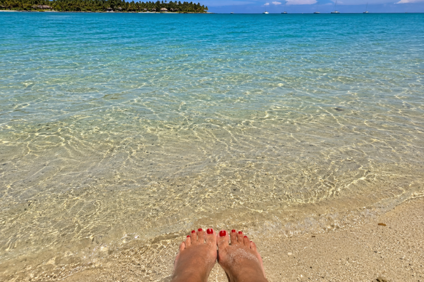 Feet resting in shallow clear water on a tropical beach with palm trees and boats in the distance.
