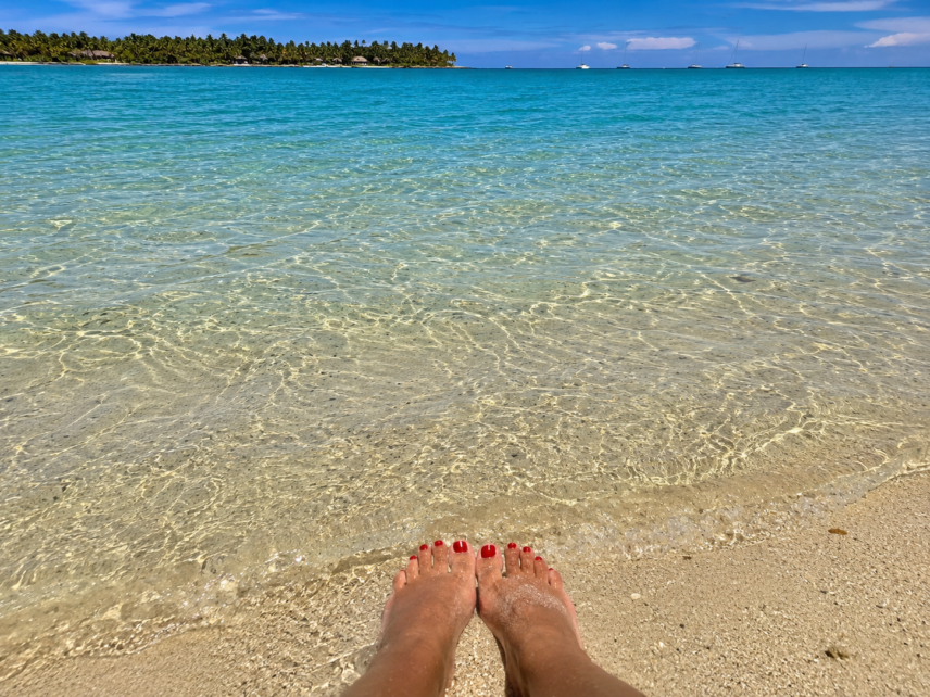 Feet resting in shallow clear water on a tropical beach with palm trees and boats in the distance.